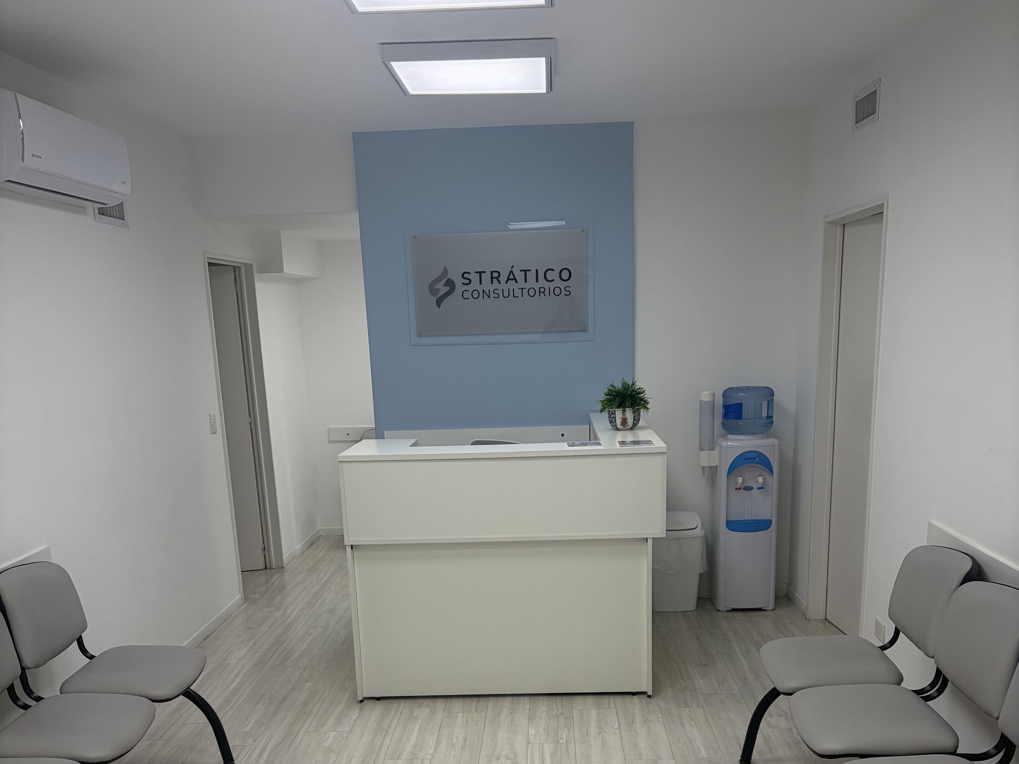 Otra vista de la recepción: el mostrador con una planta y el dispenser de agua al lado, frente a la pared celeste con el logo